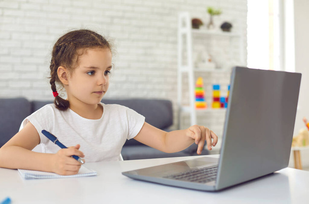 Online education. Video lesson school for children. A little girl is watching a video lesson in a laptop sitting at a table in the room. - Foto, Imagem