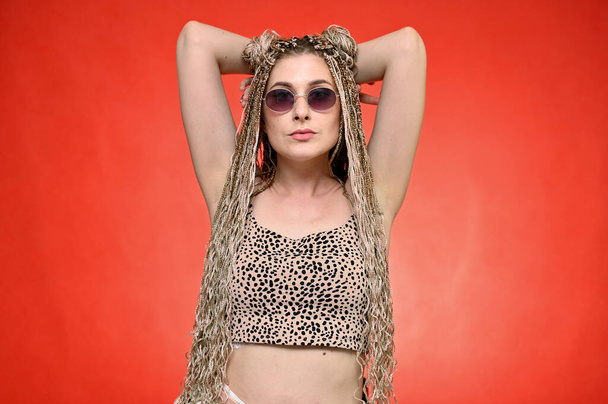 Pretty model with glasses posing on a red background standing right in front of the camera. Portrait of a caucasian fashionable woman with afro dreadlocks long hair in studio - Fotó, kép