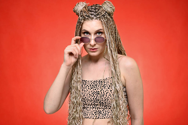 Portrait of a caucasian fashionable woman with afro dreadlocks long hair in studio. Pretty model with glasses posing on a red background standing right in front of the camera - Photo, image