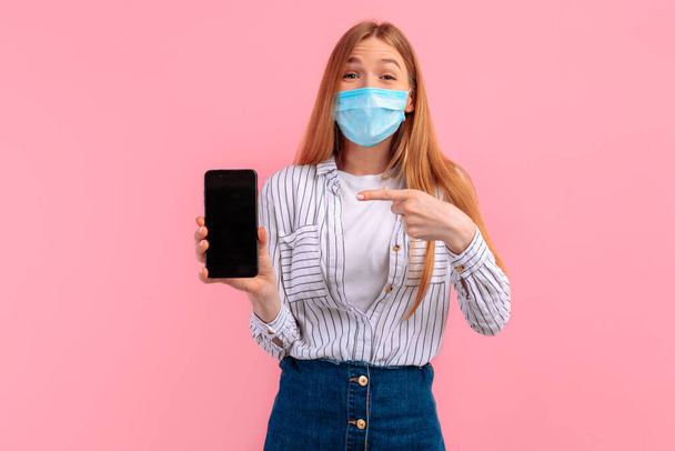 excited happy young woman wearing a medical protective face mask shows a blank mobile phone screen for a copy of the space, on an isolated pink background - Foto, Imagen