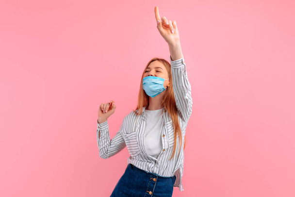 young woman with a medical protective mask on her face, dancing, active young woman having fun, on a pink background - Foto, imagen