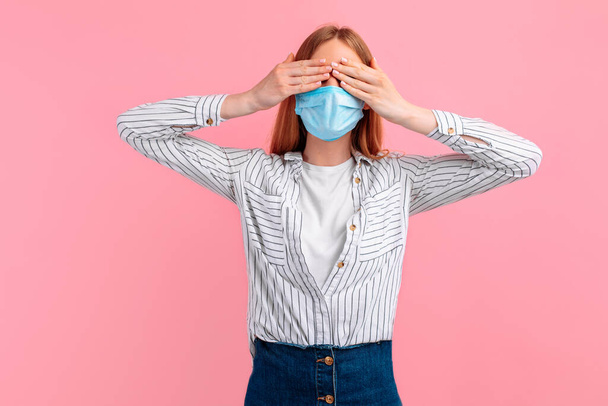 young woman in a medical protective mask covers her eyes with her hands on an isolated pink background. coronavirus - Foto, afbeelding