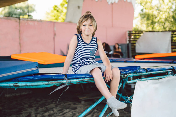 The theme of childhood, sports and health. Little boy gymnast resting in trampoline training session. A child athlete sits on a trampoline and looks at the camera. Acrobatics Sports Club for children. - Φωτογραφία, εικόνα