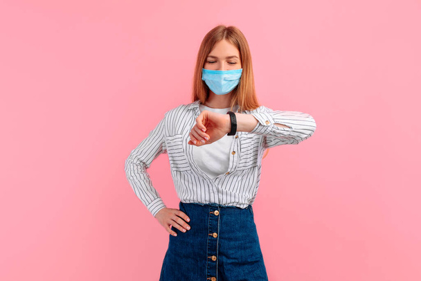 young smiling businesswoman in a medical mask, looking at the clock on her hand, on a pink background - Photo, image