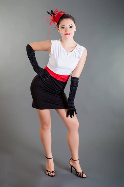 красива молода струнка дівчина в чорно-білій сукні з червоним поясом і довгими чорними рукавичками позує на сірому фоні в студії
 - Фото, зображення