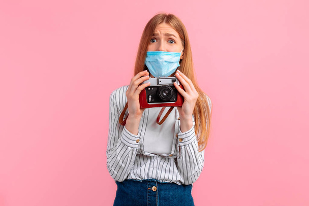 distraught, shocked young woman wearing a medical protective mask on her face, standing with a camera on a pink background. Travel, tourism, coronavirus, - Photo, image