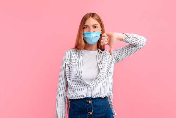 disgruntled annoyed unhappy young woman in a medical protective mask, the woman gives a thumbs down as an unfriendly gesture on a pink background - Фото, зображення