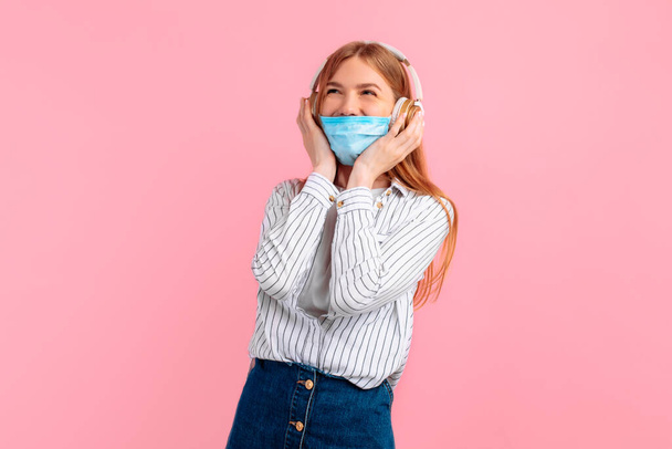 Portrait of a cheerful young woman in a medical protective mask, standing isolated on a pink background, listening to music with headphones - Фото, зображення