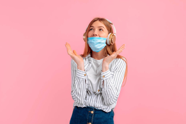 cheerful stylish young woman in a medical protective mask, standing isolated on a pink background, listening to music with headphones - Photo, image