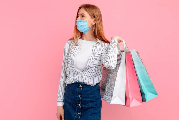 Happy girl in a medical mask on her face, posing with shopping bags, on a pink background. Shopping, coronavirus - Photo, image
