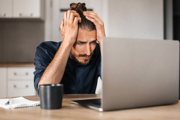 Portrait of a confused young freelancer, a man working on a laptop at home looking at a computer screen with an upset face. An upset worker in a shirt wraps his arms behind his head.  - Photo, Image
