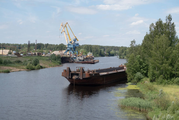 An empty bulk cargo barge moored to the shore of a small river amid cranes loading gravel - Foto, Bild