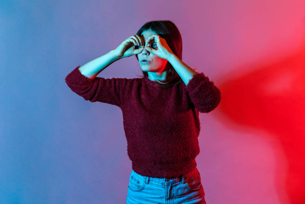 Neon light portrait of woman looking through binoculars hand gesture with attentive view, observing far distance and zooming to discover something interesting, spying. indoor studio shot isolated - Foto, immagini