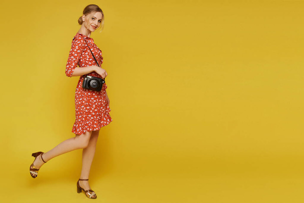 Full-length portrait of a young woman in a light dress and with a photo camera on the yellow background, isolated. Traveler concept - Photo, Image