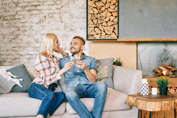 Giovane coppia felice bere champagne e sorridere seduto sul divano nella loro casa moderna - Foto, immagini