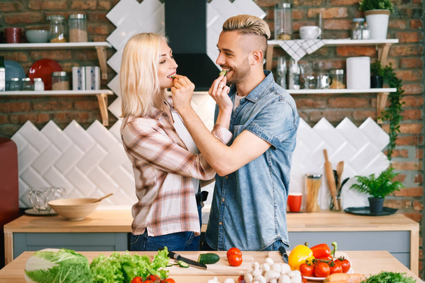 Beautiful young couple is feeding each other and smiling while cooking in kitchen at home - Фото, изображение