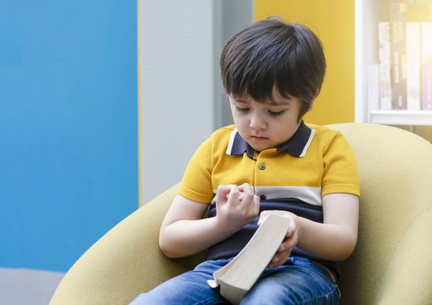 Niño solitario con la cara aburrida mirando sus dedos mientras está sentado en la tienda de libros, niño de la escuela sosteniendo un libro sentado en la silla amarilla en la biblioteca, concepto de actividad interior de los estudiantes de primer año
 - Foto, Imagen