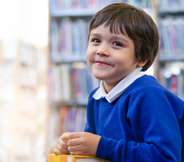 Vorschulkind genießt das Lesen eines Cartoons in der Bibliothek mit verschwommenem Hintergrund des Bücherregals, Netter Junge schaut mit lächelndem Gesicht in die Kamera, während er Lieblingsbuch in der Schulbibliothek liest, - Foto, Bild