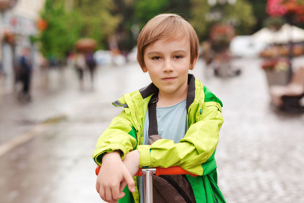 Aranyos 10 éves fiú portréja a szabadban. Jóképű kölyök sétál a város utcáján. Gyerekdivat, életstílus. Sportos fiú robogóval. A fiú a kamerába néz. Férfi frizura. Szőke hajú gyerek.. - Fotó, kép