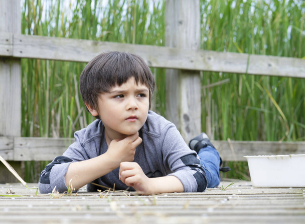 Outdoor portrait of kid boy lying on wooden bridge and looking out with thniking face, Active child having fun playing in wildlife park,Kid having adventure in nature reserve in sunny day summer - Photo, Image