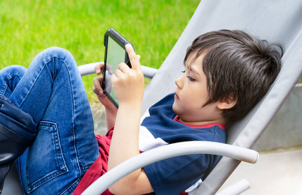 Portrait Kind sitzt auf Schaukelstuhl und spielt Spiel auf Tablet im Garten, Kind Junge hat Spaß beim Ansehen von Cartoons auf digitalem Taplet, Kind mit lächelndem Gesicht spielt Spiele auf Touchpad. - Foto, Bild