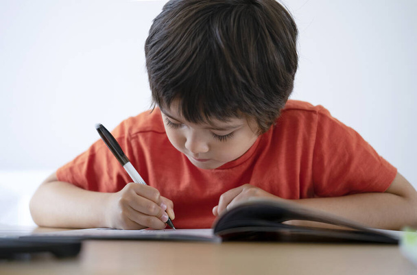 Portret chłopca z przedszkola siedzącego na stole odrabiającego lekcje z białym tłem, Happy Child trzymającego czarny długopis, Mały chłopiec pisze na białym papierze przy stole, Szkoła podstawowa i koncepcja edukacji - Zdjęcie, obraz