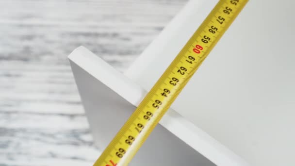 Measurement of furniture with a yellow metal measuring tape. White cabinet shelf on wooden background close-up - Πλάνα, βίντεο