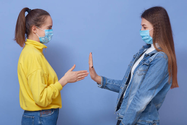 Young girl in yellow sweater wants to greet her friend shaking hands, female in denim jacket and mask refuses to protect herself from virus, posing isolated over blue wall. - Photo, image