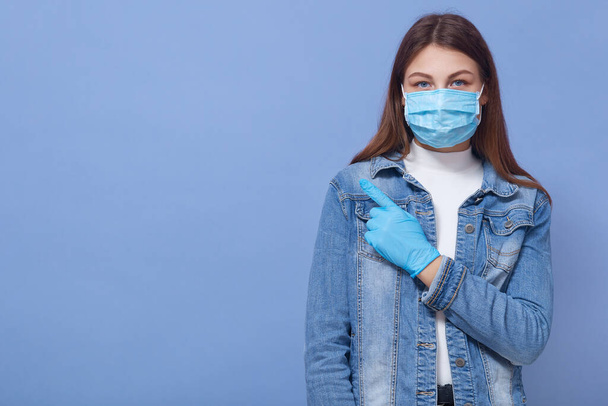 Young attractive dark haired fashionable girl wearing medical mask and latex gloves points aside with her index finger, looking at camera, poses against blue wall. - Fotoğraf, Görsel