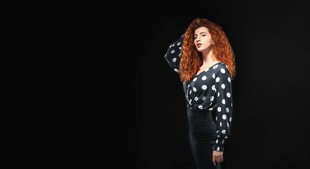 Isolated slender red-haired curly girl in a blouse and a black skirt against a black background. Office employee. - Фото, изображение