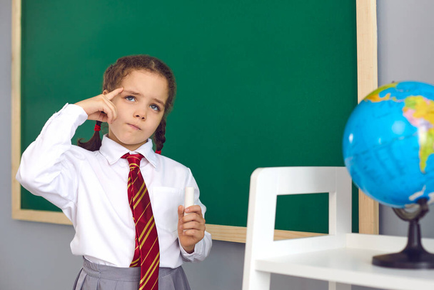 Back to school. A happy schoolgirl thinks about the task while standing at the blackboard in the classroom. - Φωτογραφία, εικόνα