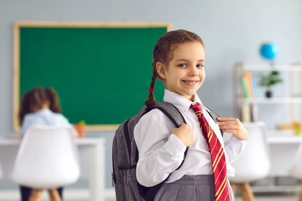 Back to school. Portrait of happy little girl with backpack and uniform standing in classroom, copy space - Foto, Imagem