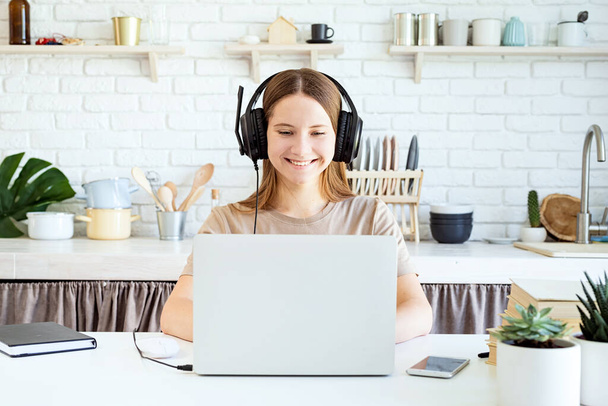 Uczenie się na odległość. Uśmiechnięta nastolatka w słuchawkach, studiująca na laptopie w domowej kuchni. - Zdjęcie, obraz