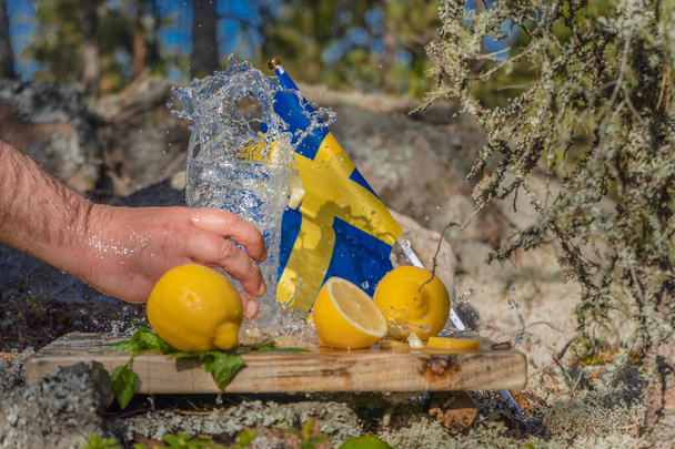 Летние закуски. Стакан воды в мужской руке. Лимоны, лаймы и мята на фоне скандинавской природы и шведского флага. Различные комбинации капель воды. Швеция
 - Фото, изображение