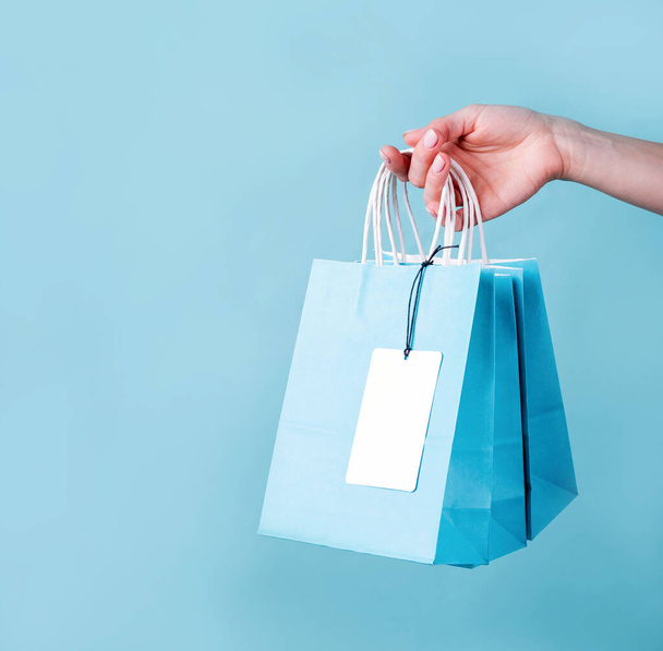 woman hand carrying a bunch of blue shopping bags with a blank tag over blue background with copy space - Фото, зображення