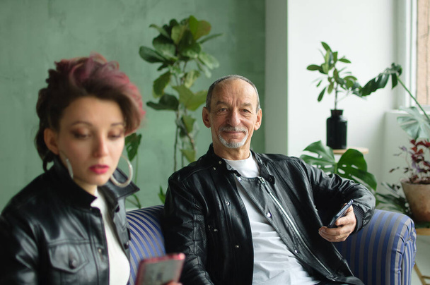 Family portrait of adult daughter and senior father in loft room with houseplants. Man and girl are wearing black leather jackets in punk style and browsing on their smartphones without communication - Фото, изображение