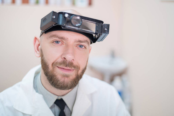 Male bearded doctor in a medical coat and with a magnifying head strap w lights sitting on the desk. Optometrist equipment. Medical office. - Foto, imagen