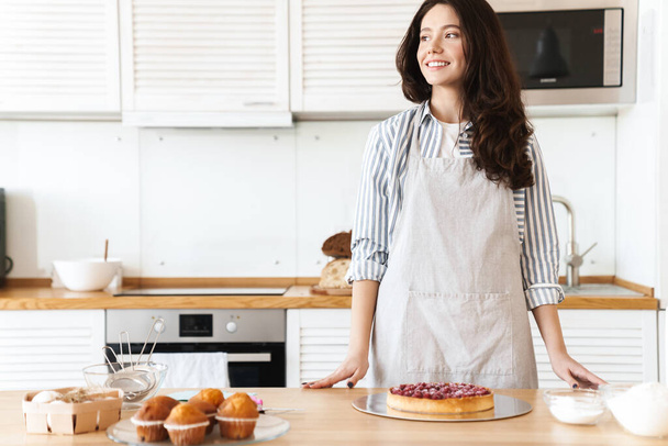 Image of pleased brunette woman wearing apron smiling while cooking pie in modern kitchen - Foto, Bild