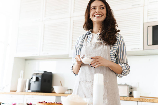 Image of joyful brunette woman wearing apron smiling while cooking pie in modern kitchen - Photo, image