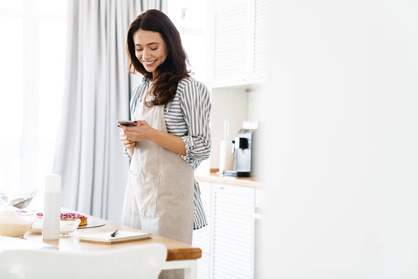 Image of smiling brunette woman wearing apron using cellphone while cooking pie in modern kitchen - Foto, Imagen