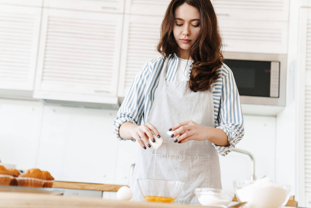 Image of thinking brunette woman wearing apron preparing dough while cooking pie in modern kitchen - Zdjęcie, obraz