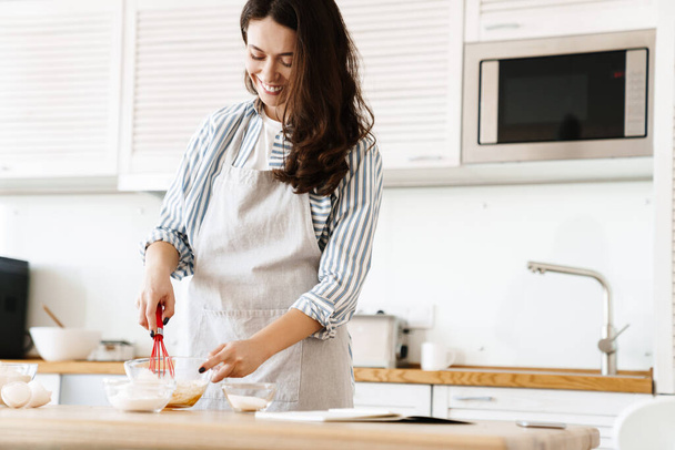 Image of cheerful brunette woman wearing apron preparing dough while cooking pie in modern kitchen - 写真・画像