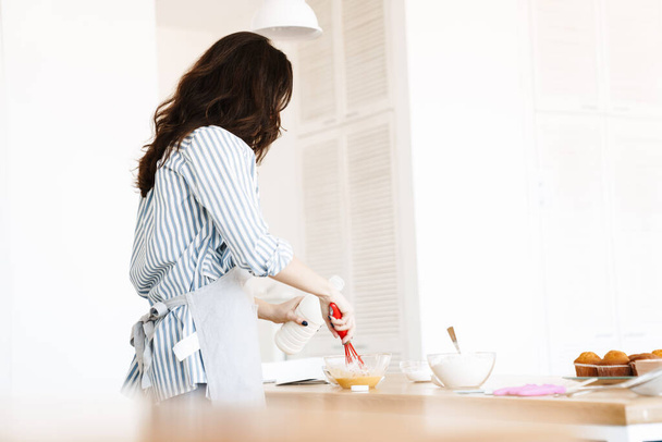 Image of caucasian brunette woman wearing apron preparing dough while cooking pie in modern kitchen - Fotografie, Obrázek