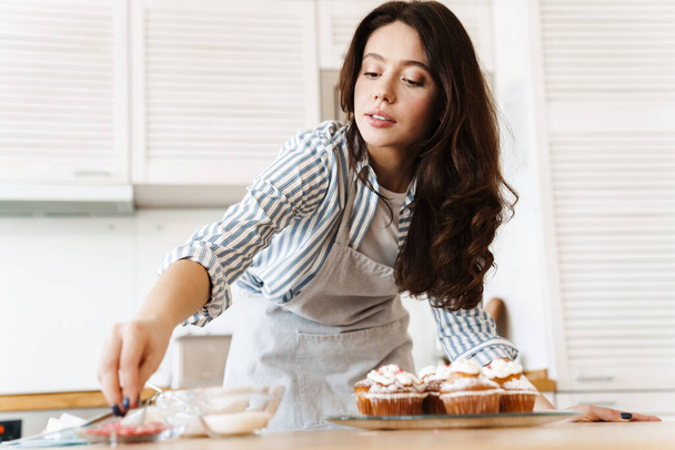 Image of caucasian concentrated woman wearing apron cooking muffins in modern kitchen - Foto, immagini