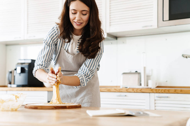 Image of caucasian smiling woman wearing apron kneading dough for sweet pie in modern kitchen - Фото, изображение