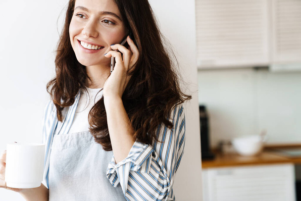 Image of happy beautiful woman wearing apron talking on cellphone while drinking coffee in modern kitchen - Фото, зображення