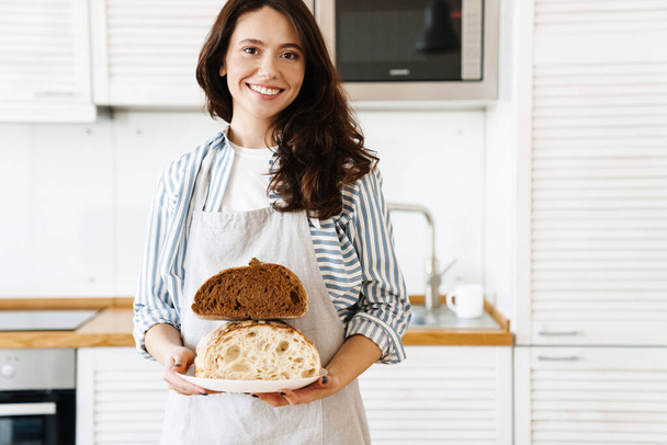 Obrázek šťastné krásné ženy nosí zástěru s úsměvem, zatímco ukazuje chléb na kameru v moderní kuchyni - Fotografie, Obrázek