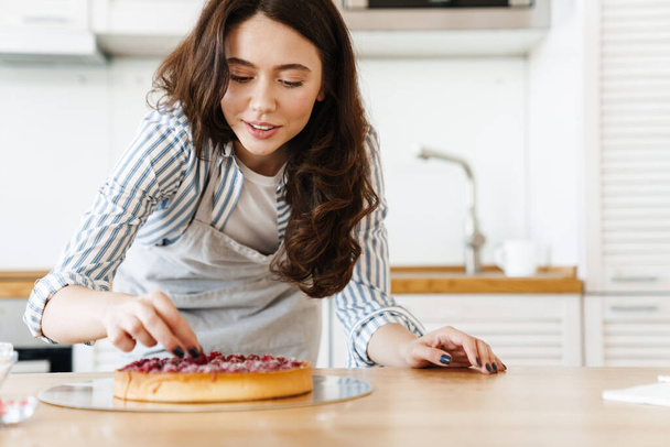 Image of pleased beautiful woman wearing apron cooking pie with raspberries in modern kitchen - Foto, Imagem