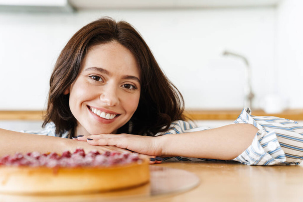 Image of pleased beautiful woman smiling and cooking pie with raspberries in modern kitchen - Fotografie, Obrázek
