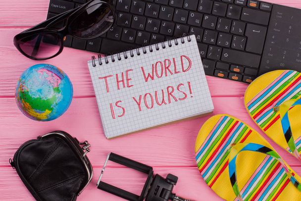 De wereld is van jou op notebook met vrouwen reiziger accessoires bril portemonnee en teenslippers op roze tafel top achtergrond. - Foto, afbeelding